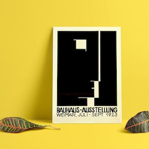 May include: A framed Bauhaus-Ausstellung poster with a black and white geometric design. The poster includes the text "BAUHAUS-AUSSTELLUNG WEIMAR, JULI-SEPT. 1923" and "living room wall art" on a yellow background. Two leaves are placed in front of the poster.