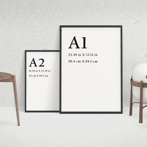 May include: Two framed art prints with black borders, labeled A1 and A2, with dimensions in inches and centimeters. A wooden chair and a decorative plant are on the left. A white sphere on a wooden stand and a vase with greenery are on the right.