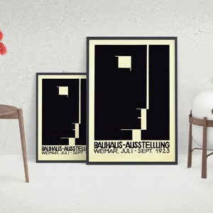 May include: Two framed Bauhaus-Ausstellung posters with black and white geometric designs. The posters are displayed with a wooden chair holding a white vase with red flowers, and a small potted plant. A white sphere sits on a wooden stand.