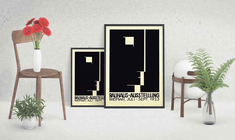 May include: Two framed Bauhaus-Ausstellung posters with black and white geometric designs. The posters are displayed with a wooden chair holding a white vase with red flowers, and a small potted plant. A white sphere sits on a wooden stand.