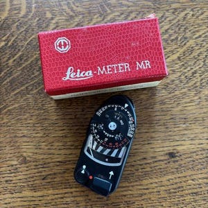 May include: A vintage Leica-Meter MR light meter with a black body and a red box. The box is red with the words "Leica-METER MR" printed on it. The light meter has a dial with numbers and markings.