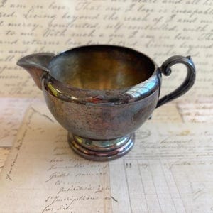 May include: Antique silver-plated creamer with a spout and handle. The creamer has a round body and sits on a pedestal base. The interior is a shiny silver color, and the exterior has a tarnished, vintage appearance.