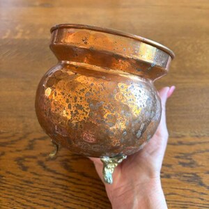May include: A copper cauldron with a rounded body and three ornate feet. The cauldron has a hammered texture and a tarnished patina, with a wide, slightly angled rim. The overall color is a mix of copper and bronze.