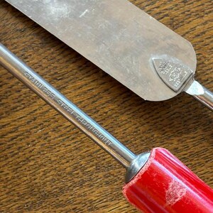 May include: Close-up of two stainless steel kitchen tools. One is a spatula with a silver blade and a red handle. The other tool has a red handle and the text "STAINLESS STEEL THROUGHOUT" printed on the metal. The tools are on a wooden surface.