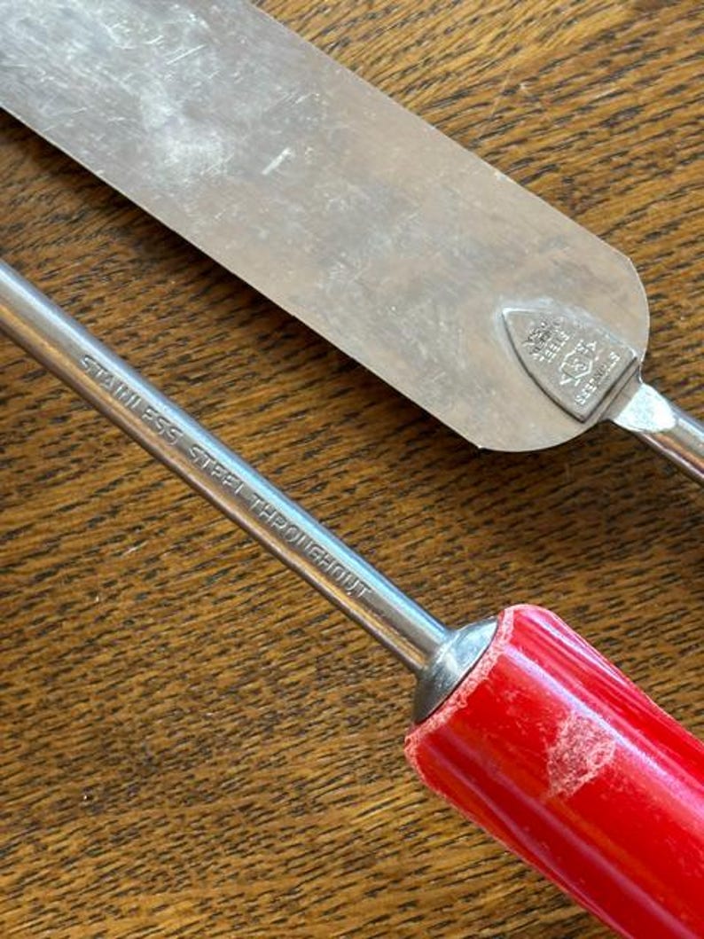 May include: Close-up of two stainless steel kitchen tools. One is a spatula with a silver blade and a red handle. The other tool has a red handle and the text "STAINLESS STEEL THROUGHOUT" printed on the metal. The tools are on a wooden surface.