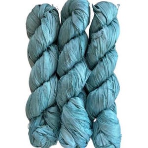 May include: Three skeins of teal blue ribbon yarn arranged vertically. The yarn has a flat, crinkled, and textured appearance with a matte finish, wound into long, twisted bundles that show consistent color throughout the material.
