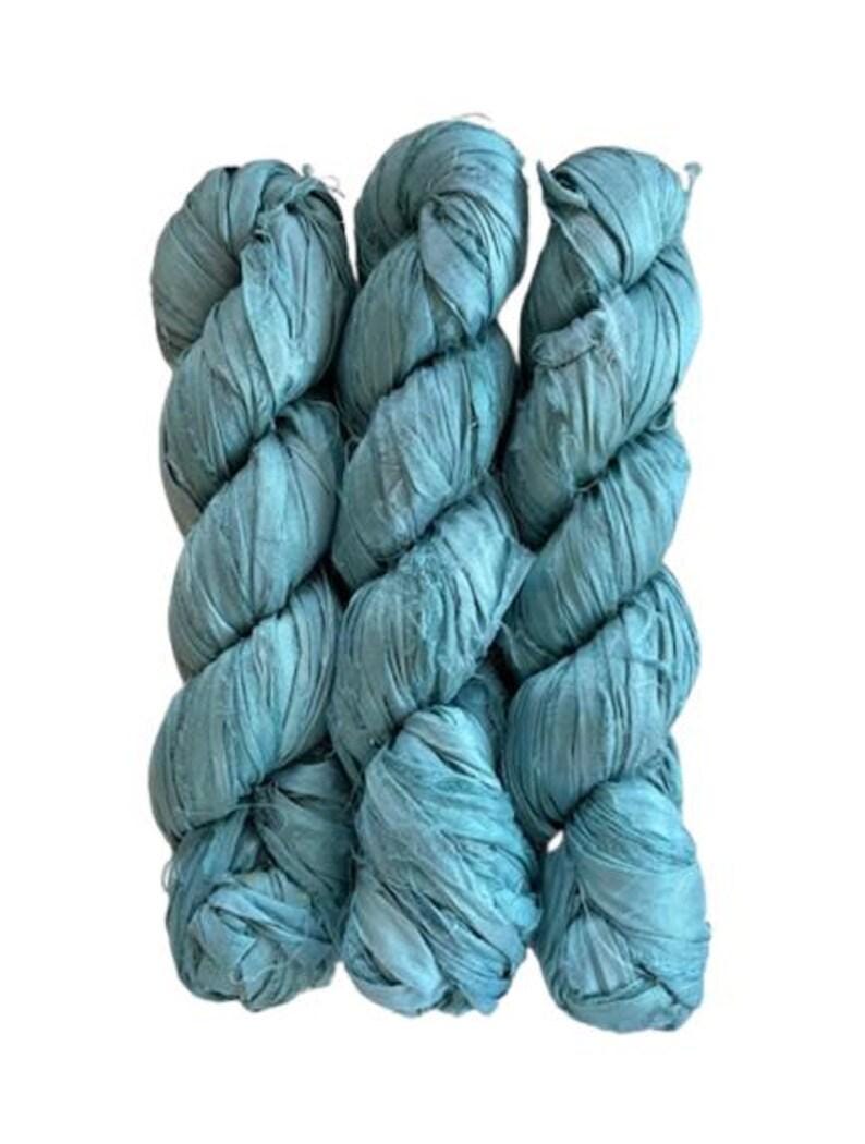 May include: Three skeins of teal blue ribbon yarn arranged vertically. The yarn has a flat, crinkled, and textured appearance with a matte finish, wound into long, twisted bundles that show consistent color throughout the material.