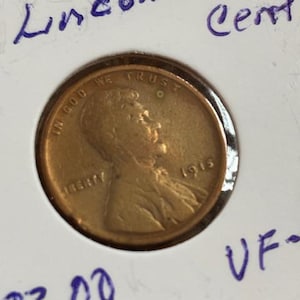 May include: A copper-colored 1915 Lincoln Wheat Cent coin, displayed in a protective holder. The coin features a profile of Abraham Lincoln and the words "IN GOD WE TRUST" and "LIBERTY". The holder has handwritten text including "Lincoln Wheat Cent".
