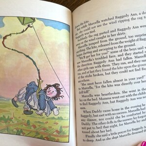 Puede incluir: Una ilustraci&oacute;n vintage de una mu&ntilde;eca Raggedy Ann volando una cometa. La mu&ntilde;eca lleva un vestido azul y un lazo amarillo. La cometa es una simple forma cuadrada con una cuerda roja, verde y amarilla. El fondo es un cielo azul claro con nubes blancas.