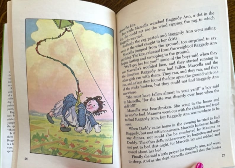 Puede incluir: Una ilustraci&oacute;n vintage de una mu&ntilde;eca Raggedy Ann volando una cometa. La mu&ntilde;eca lleva un vestido azul y un lazo amarillo. La cometa es una simple forma cuadrada con una cuerda roja, verde y amarilla. El fondo es un cielo azul claro con nubes blancas.