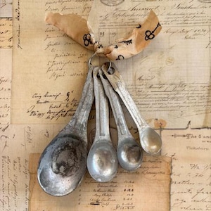 May include: A set of four vintage metal measuring spoons hanging from a tan ribbon printed with black keys. The spoons are attached to a metal ring. The background is a collage of aged paper with handwritten script.