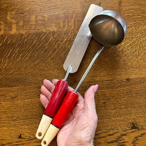 May include: Two vintage kitchen utensils: a ladle and a spatula. The ladle has a silver bowl and a red handle with a cream-colored base. The spatula is silver with a red handle and cream-colored base. The utensils are displayed on a wooden surface.