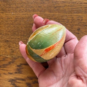 May include: A hand-painted, wooden egg-shaped decorative object. The egg features a design with green, orange, and tan colors. The object has a smooth, varnished finish and is held in a person's hand.