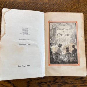 May include: An open book with a white cover and a black and white illustration of a group of people sitting around a table. The book's title is "The Story of Crisco".