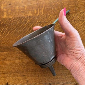 May include: A vintage metal funnel with a wide mouth and a narrow spout. The funnel is a dark, aged gray color with visible wear and tear. The funnel is held by a hand with pink nail polish. The funnel is likely used for pouring liquids.