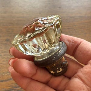 May include: A clear, faceted glass bottle stopper with a dark brown base. The glass has a ribbed design and a rounded top. The base shows signs of wear, with chipped paint. The stopper is held in a hand.