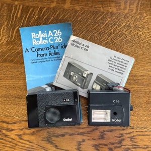 May include: Two black and silver Rollei A 26 and C 26 cameras with flash units. The cameras are on a wooden surface. The text on the packaging reads "A "Camera-Plus" idea from Rollei." and "In der Praxis In practical use dans la pratique nell'uso pratico en la práctica in de praktijk"
