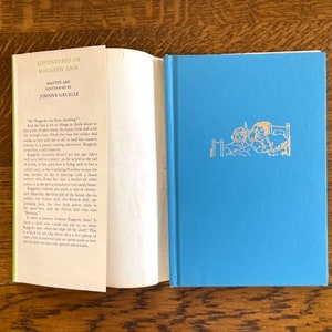 Puede incluir: Una cubierta de libro azul con el t&iacute;tulo "Adventures of Raggedy Ann" escrito en letras doradas. El libro es de Johnny Gruelle. La contraportada tiene una imagen de dos mu&ntilde;ecas en una cama.