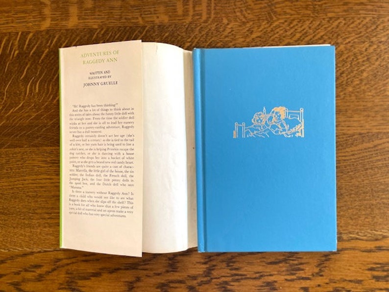 Puede incluir: Una cubierta de libro azul con el t&iacute;tulo "Adventures of Raggedy Ann" escrito en letras doradas. El libro es de Johnny Gruelle. La contraportada tiene una imagen de dos mu&ntilde;ecas en una cama.