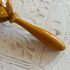 May include: A wooden maraca featuring a rounded head with a polished finish and a geometric diamond pattern in alternating shades of light and dark brown. The instrument includes a smooth, tapered handle in a solid golden-yellow wood tone.