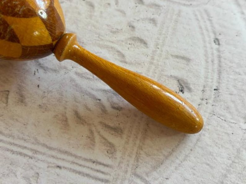 May include: A wooden maraca featuring a rounded head with a polished finish and a geometric diamond pattern in alternating shades of light and dark brown. The instrument includes a smooth, tapered handle in a solid golden-yellow wood tone.