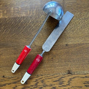 May include: A stainless steel ladle and spatula set with red handles and white tips. The ladle has a rounded bowl, and the spatula is rectangular. The set is designed for kitchen use.