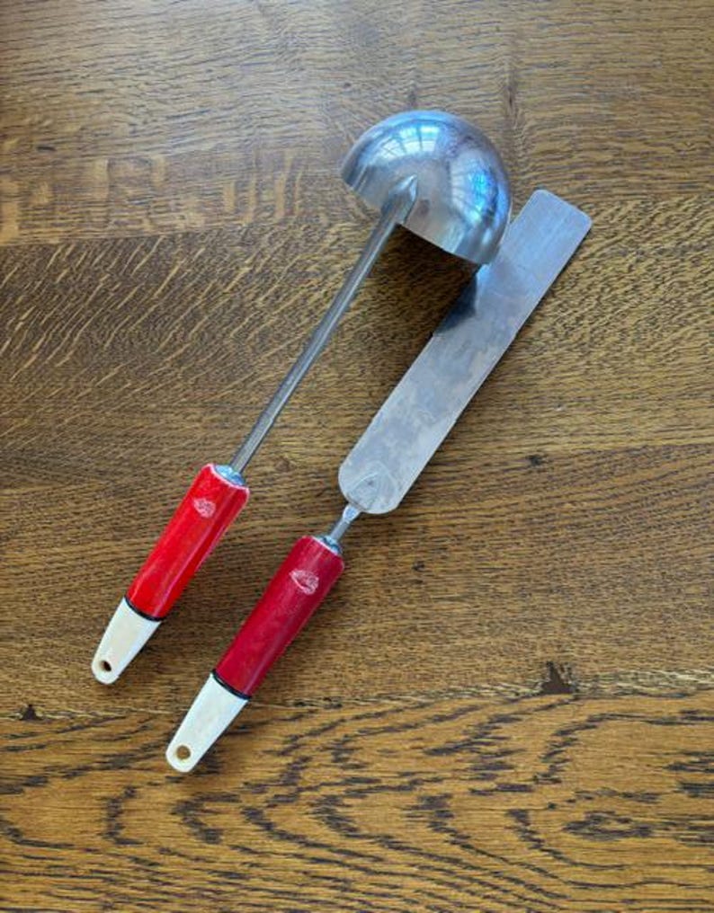 May include: A stainless steel ladle and spatula set with red handles and white tips. The ladle has a rounded bowl, and the spatula is rectangular. The set is designed for kitchen use.