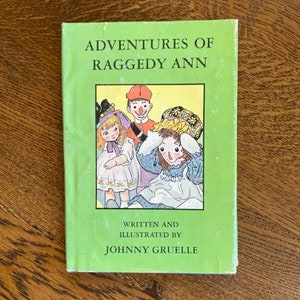 Puede incluir: Una cubierta de libro verde con el t&iacute;tulo "Adventures of Raggedy Ann" en texto blanco. La cubierta presenta una ilustraci&oacute;n de Raggedy Ann y dos mu&ntilde;ecas m&aacute;s. El texto "Written and Illustrated by Johnny Gruelle" est&aacute; impreso debajo de la ilustraci&oacute;n.