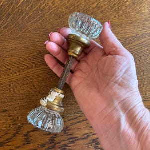 May include: A pair of clear, fluted glass door knobs connected by a central metal spindle. The knobs feature brass-colored bases, with one base showing visible white paint wear. The glass components have a faceted, circular design.