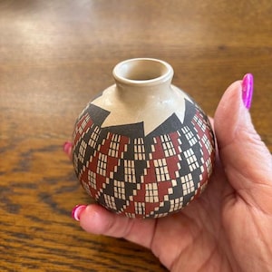 May include: A small, round ceramic vase with a cream-colored neck and a patterned body. The body features a geometric design in black, red, and white. The vase is held in a hand.