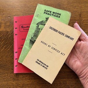 May include: Three vintage booklets on a wooden surface. The booklets are red, green, and tan. The green booklet has the text "SAFE WORK PRACTICES" and an illustration of a train. The tan booklet reads "SOUTHERN PACIFIC COMPANY HOURS OF SERVICE ACT".