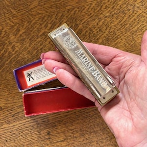 May include: A silver-toned Marine Band harmonica held in a hand, with the text "MARINE BAND M. Hohner" visible. The harmonica is next to a red and blue box with text and a graphic.