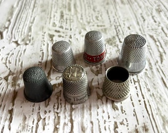 May include: A collection of six metal sewing thimbles with dimpled surfaces. One thimble features a red band with the text TOM, another displays a circular emblem with a building illustration and the text Lake Thimble on the base.