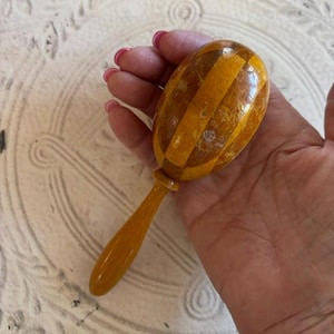 May include: A handheld wooden maraca featuring a rounded, egg-shaped head with alternating vertical stripes of light yellow and dark amber wood grain. The instrument includes a smooth, solid yellow handle attached to the base of the head.