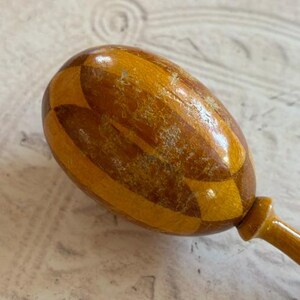 May include: A wooden maraca featuring a polished, egg-shaped head with a geometric, alternating light and dark brown wood grain pattern. The instrument has a smooth, rounded handle extending from the base of the head, showing a glossy finish.