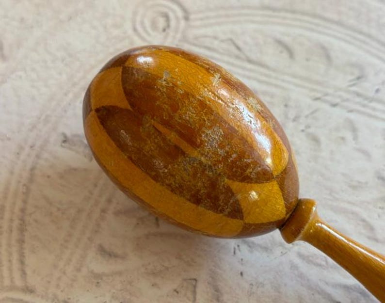 May include: A wooden maraca featuring a polished, egg-shaped head with a geometric, alternating light and dark brown wood grain pattern. The instrument has a smooth, rounded handle extending from the base of the head, showing a glossy finish.
