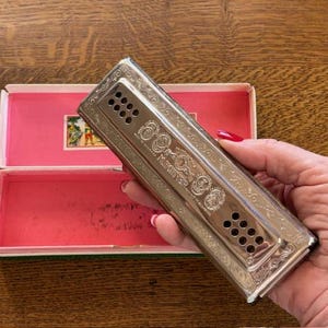 May include: A vintage silver harmonica with decorative engravings, held near its open pink and green box. The harmonica has multiple rows of holes and is labeled "M. Hohner". The box has a small picture on the inside.