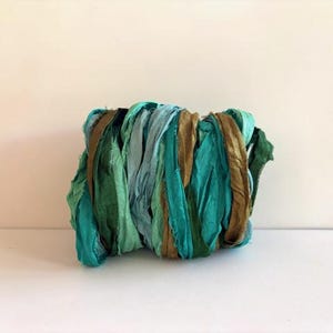 May include: A bundle of crinkled silk ribbon featuring a color palette of teal, emerald green, light blue, and metallic bronze. The fabric strips are wound together in a dense, textured cluster, showcasing a variety of shades and a soft sheen.