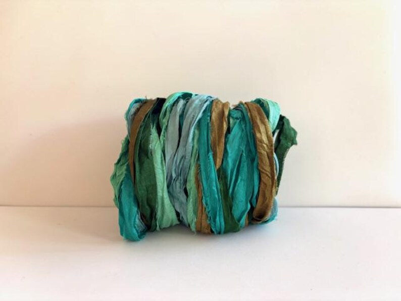 May include: A bundle of crinkled silk ribbon featuring a color palette of teal, emerald green, light blue, and metallic bronze. The fabric strips are wound together in a dense, textured cluster, showcasing a variety of shades and a soft sheen.