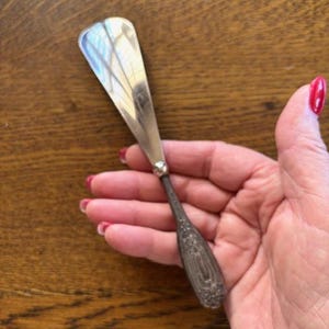 May include: A silver butter spreader with a decorative handle. The blade is wide and flat, reflecting light. The handle features an ornate floral design. This vintage utensil is likely used for serving butter or other spreads.