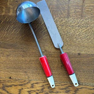 May include: A stainless steel ladle and spatula set with red handles and white tips. The ladle has a round bowl, and the spatula is rectangular. The set is designed for kitchen use.