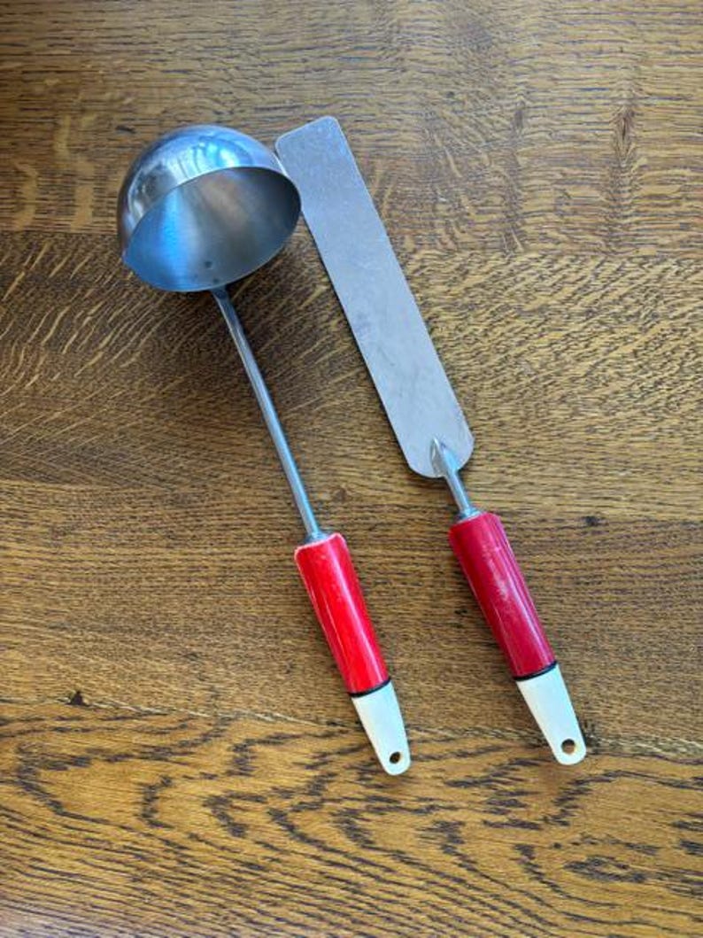May include: A stainless steel ladle and spatula set with red handles and white tips. The ladle has a round bowl, and the spatula is rectangular. The set is designed for kitchen use.