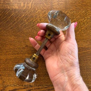 May include: A pair of clear, faceted glass door knobs connected by a central metal spindle. Each knob features a rounded, multi-faceted design attached to a circular, weathered brass-toned metal base with a visible screw hole for mounting.