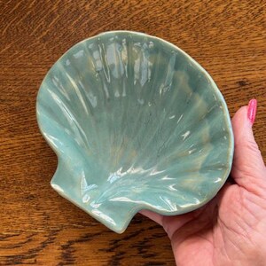 May include: A turquoise ceramic seashell-shaped dish. The dish has a glossy finish and is held in a hand. The dish is a light blue-green color with subtle variations in tone, suggesting a handmade quality. The dish is likely used for decorative purposes.