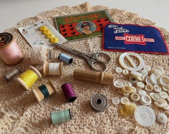 May include: A collection of vintage sewing supplies including wooden spools of thread in pink, yellow, green, purple, and light blue, metal scissors, a metal bobbin, various white buttons, and packaging labeled Starnap and Cornet.