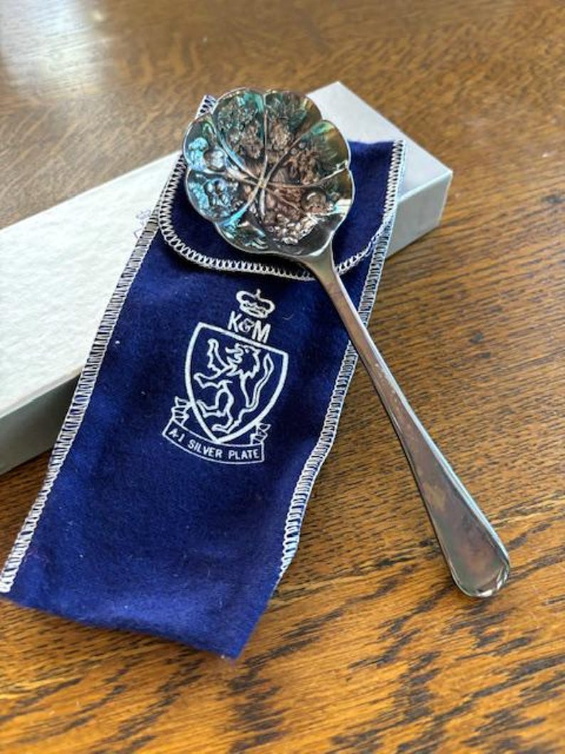Puede incluir: Una cuchara de plata con un cuenco en forma de hoja y una bolsa de terciopelo azul. La bolsa tiene un escudo con las letras "KGM" y las palabras "A1 Silver Plate".