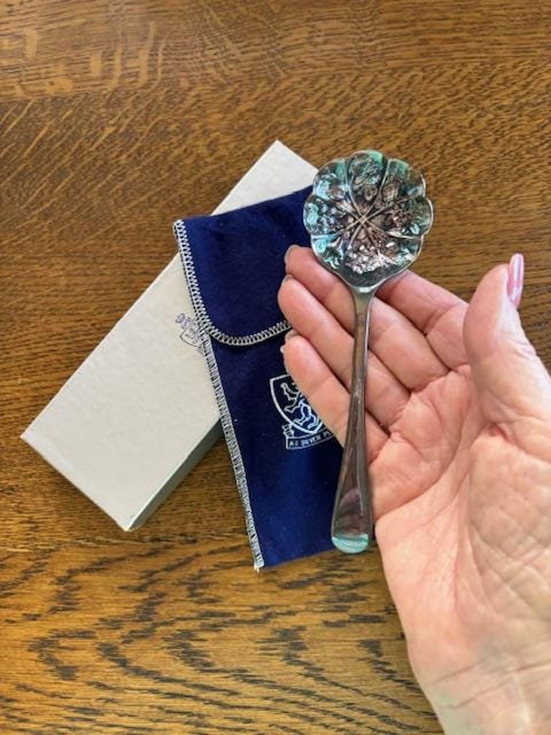 Puede incluir: Una cuchara de plata con un cuenco en forma de flor y un dise&ntilde;o detallado. La cuchara est&aacute; en una bolsa de terciopelo azul con un escudo que dice "At John's Place".
