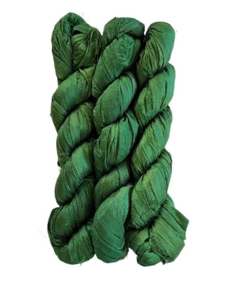 May include: A bundle of emerald green silk ribbon yarn featuring a crinkled, textured surface. The material is wound into a long, twisted skein with frayed edges visible along the length of the fabric strips, showcasing a consistent deep green hue.