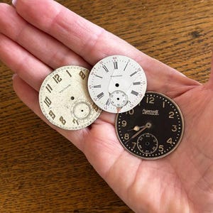 May include: Three circular watch dials including a cream-colored Waltham dial with Arabic numerals, a white Waltham dial with Roman numerals and a small crack, and a black Ingersoll Waterbury dial with Arabic numerals and a single metal hand.