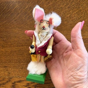 May include: A wooden rabbit figurine with a white fur head, pink ears, and small round glasses. The rabbit wears a burgundy vest with a white lace collar and holds a black and gold accessory. It stands on a green base.